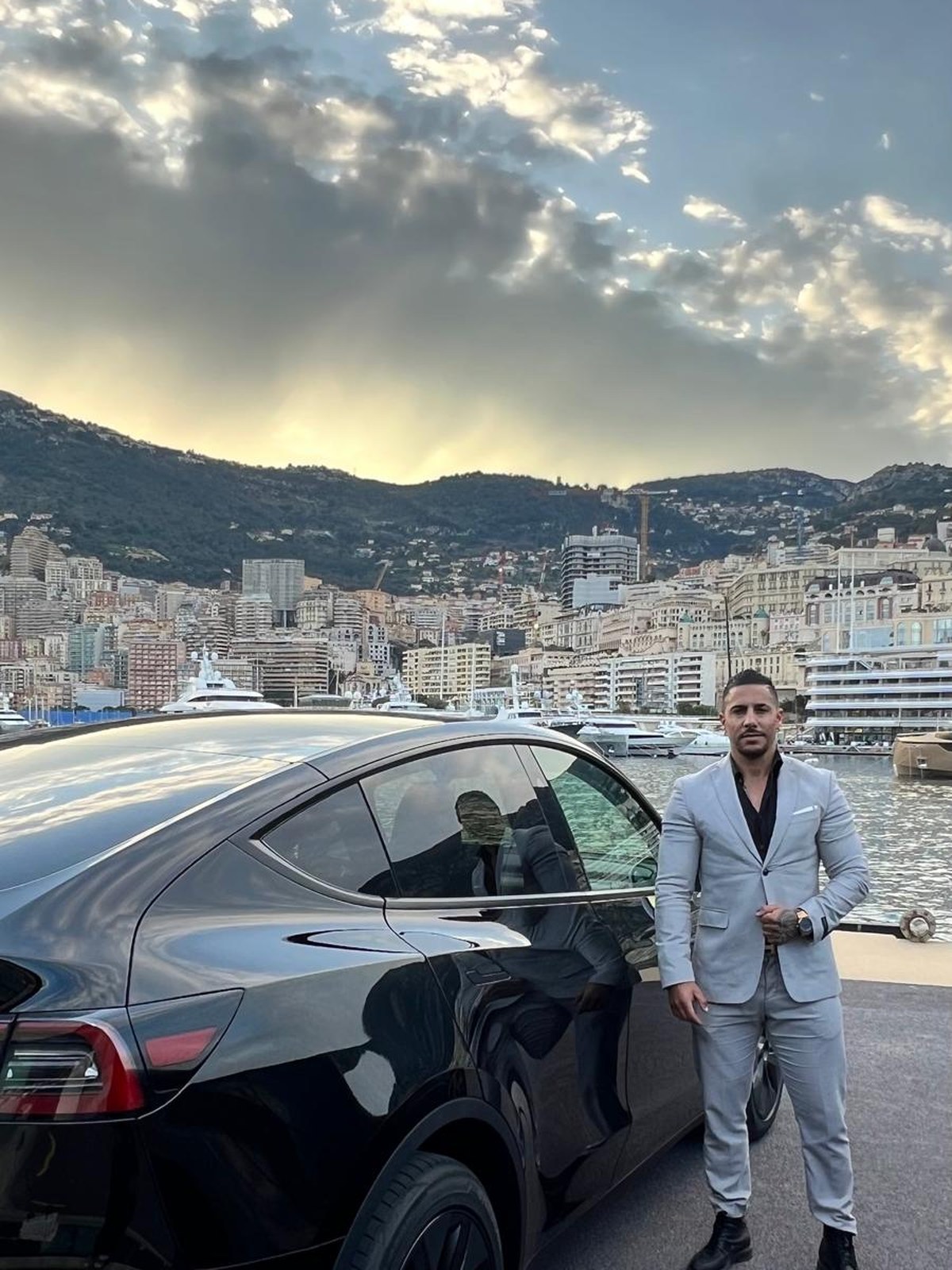 Helder Costa en costume, debout devant sa Tesla, le port de Monaco derrière lui